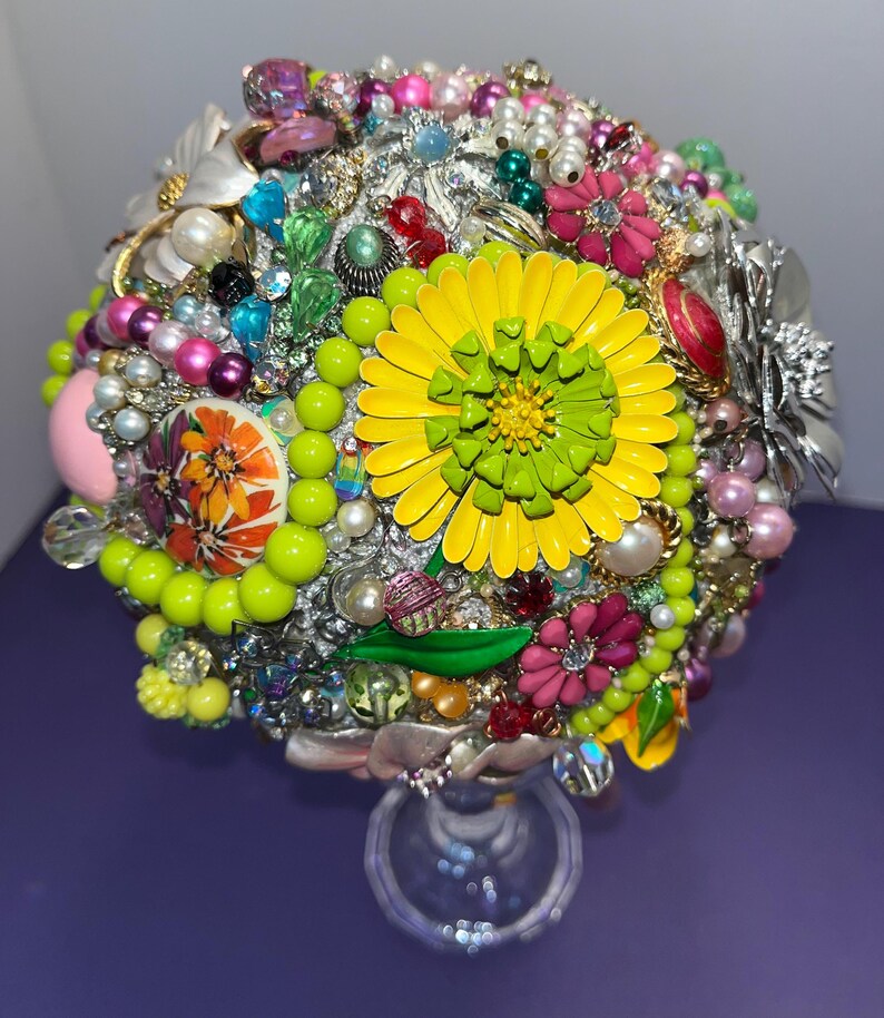 May include: A colorful bouquet made of vintage brooches and jewelry, featuring a large yellow flower with a green center. The bouquet is arranged on a clear glass stand.