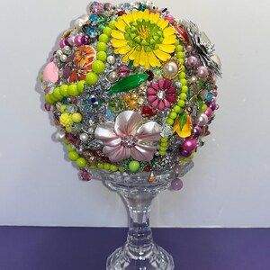 May include: A colorful sphere covered in various jewelry and trinkets, including flowers, beads, and rhinestones. The sphere is displayed on a clear glass pedestal.