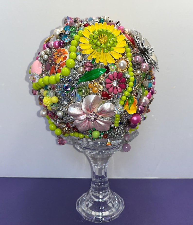 May include: A colorful sphere covered in various jewelry and trinkets, including flowers, beads, and rhinestones. The sphere is displayed on a clear glass pedestal.