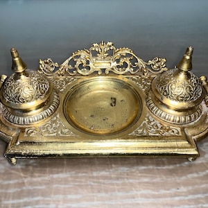 Könnte beinhalten: Ein goldfarbener Tintenfässchen aus Metall mit zwei verzierten Deckeln und einer zentralen Vertiefung. Das Tintenfässchen hat komplizierte florale Designs und einen dekorativen Rand.