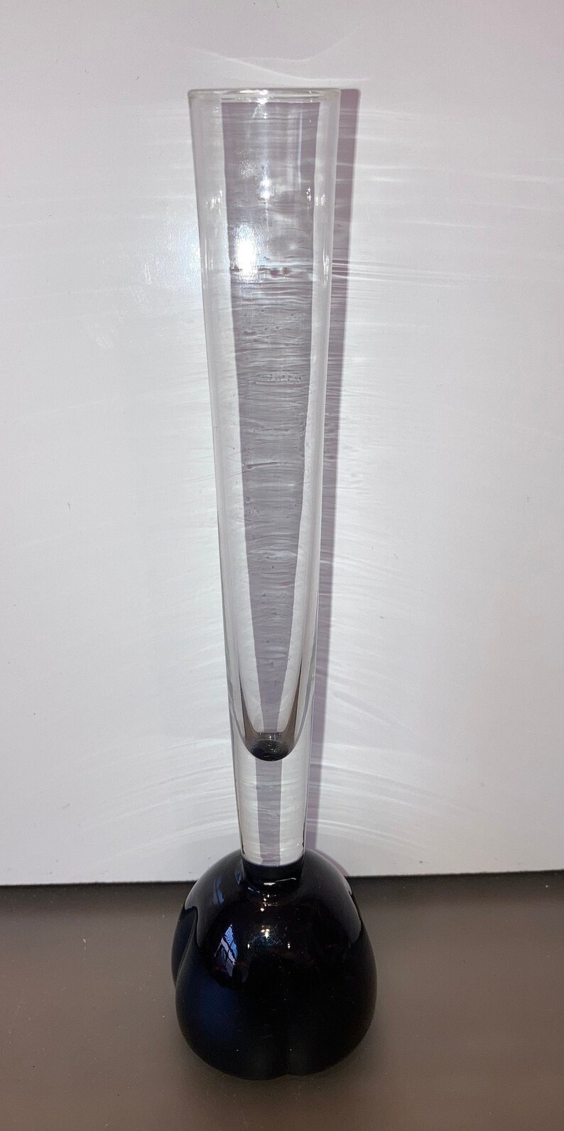 May include: A clear glass bud vase with a black, rounded base. The vase is tall and slender, perfect for displaying a single flower.