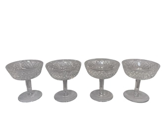 May include: Four clear, stemmed champagne coupe glasses. Each glass has a wide, shallow bowl with a diamond-cut pattern and a short stem. The glasses are arranged in a row against a white background.