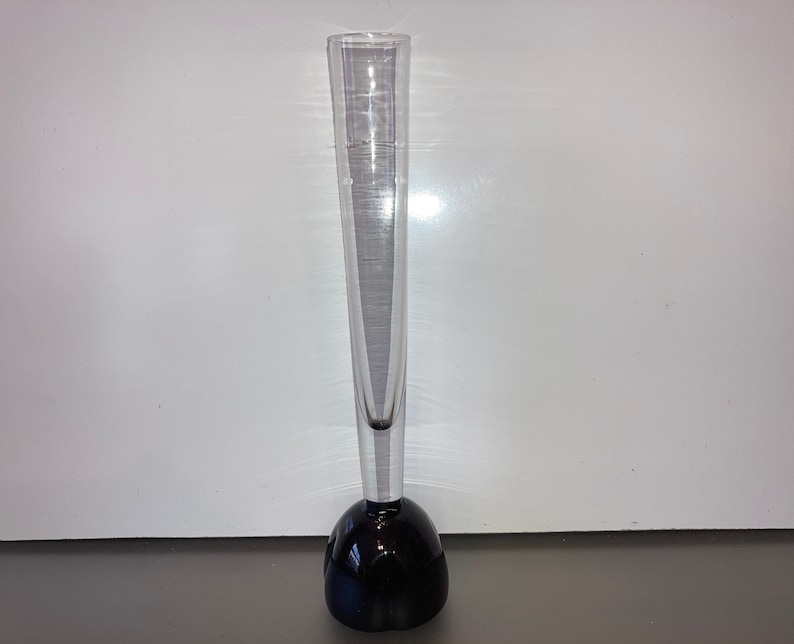May include: A clear glass vase with a tall, slender body and a round, black base.