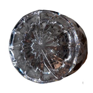 May include: A clear, faceted glass object with a circular base and a textured, geometric pattern. The glass reflects light, creating a sparkling effect. The object appears to be a decorative piece or a part of a larger item.