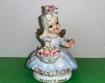 May include: A porcelain figurine of a girl with blonde hair wearing a blue dress and holding a basket of pink flowers. The figurine is titled "Friday's Child".