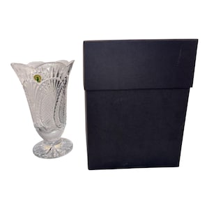 May include: A clear, cut-glass vase with a scalloped rim and a pedestal base, featuring intricate fan and diamond patterns. A small, circular green sticker with a gold seahorse logo is attached to the upper side. Beside it sits a plain black box.