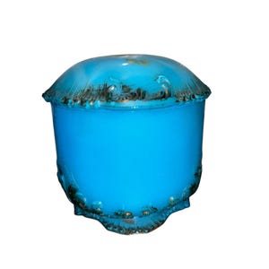 May include: A turquoise-blue, lidded jar with a rounded top and a cylindrical body. The rim and base feature a decorative, dark-colored trim. The jar has a glossy finish and is set against a white background.