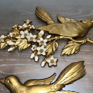 Pode incluir: Duas esculturas de parede de p&aacute;ssaros em metal dourado. Cada p&aacute;ssaro est&aacute; empoleirado em um galho com folhas douradas e flores brancas e douradas. Os p&aacute;ssaros t&ecirc;m desenhos detalhados de penas e asas.
