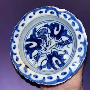 May include: A small, white ceramic dish with a blue and white floral design. The dish has a scalloped edge and a blue rim.