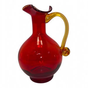 May include: A vibrant red glass pitcher featuring a rounded body, a narrow neck with a flared pouring spout, and a contrasting amber-coloured glass handle that curves gracefully from the neck to the base, ending in a small decorative glass swirl.
