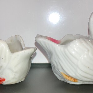 May include: Two white ceramic swan-shaped planters with painted details. One planter has a pink rim and orange accents, the other has red accents.