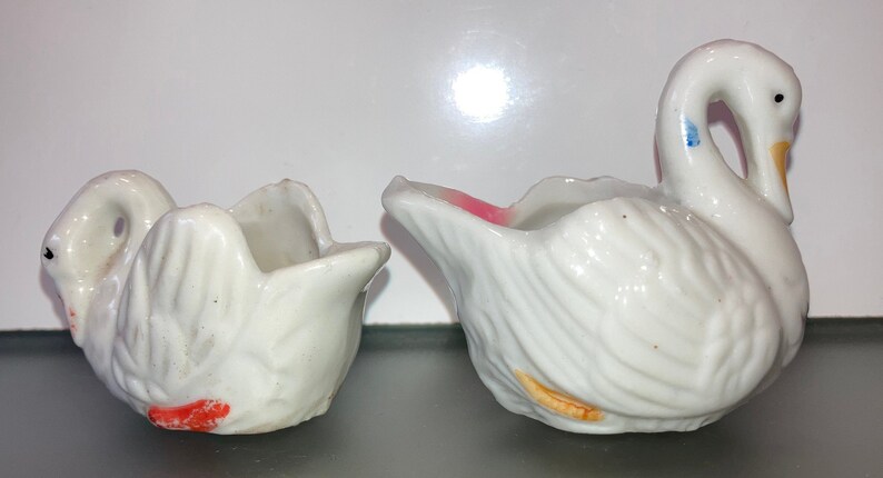 May include: Two white ceramic swan-shaped planters with painted details. One planter has a pink rim and orange accents, the other has red accents.