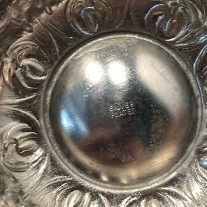 May include: Close-up of a silver-plated decorative object. The object has an ornate, floral design around the edge. The center of the object has the words "SILVER PLATED" stamped into the metal.