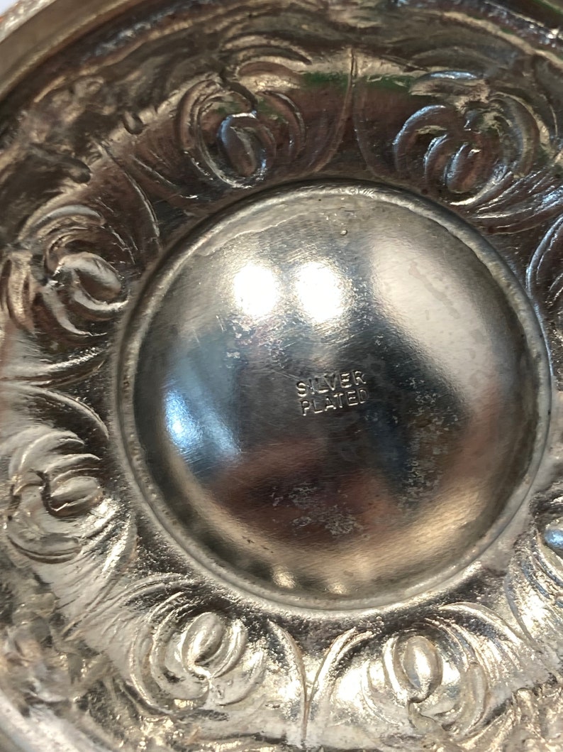 May include: Close-up of a silver-plated decorative object. The object has an ornate, floral design around the edge. The center of the object has the words "SILVER PLATED" stamped into the metal.