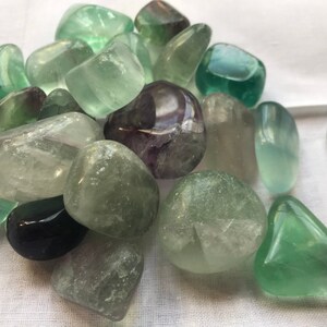 May include: A collection of green and purple fluorite tumbled stones. The stones are smooth and polished, with a variety of shades of green and purple.