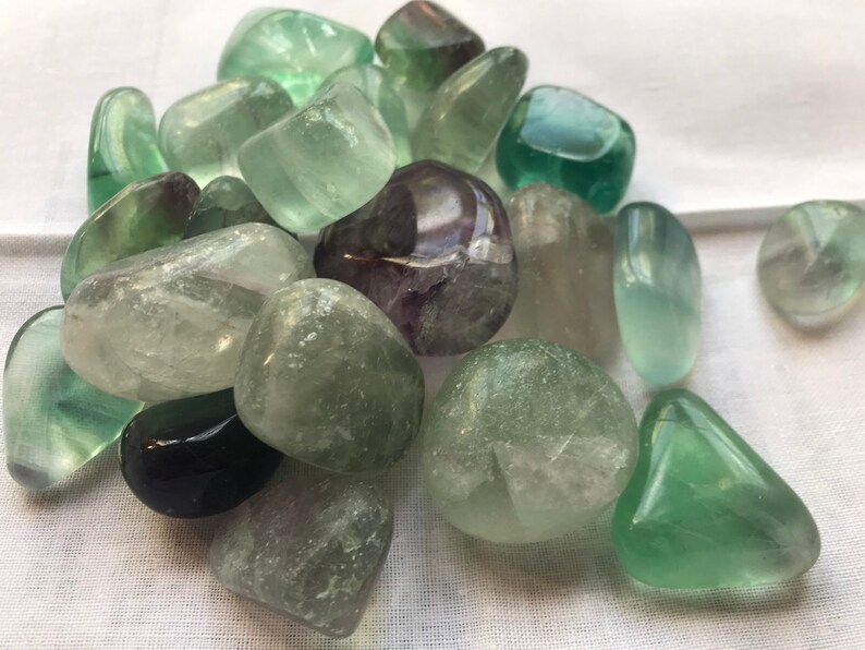 May include: A collection of green and purple fluorite tumbled stones. The stones are smooth and polished, with a variety of shades of green and purple.