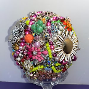 May include: A colorful bouquet made of vintage brooches, rhinestones, and pearls. The bouquet is arranged in a round shape and is held together by a clear, glass vase.
