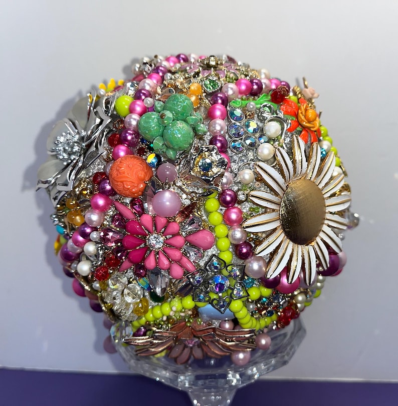 May include: A colorful bouquet made of vintage brooches, rhinestones, and pearls. The bouquet is arranged in a round shape and is held together by a clear, glass vase.