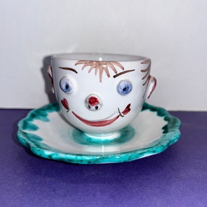 May include: A quirky white ceramic teacup and saucer set. The teacup is decorated with a smiling face, including blue eyes, a red nose, and a red smile. The saucer has a scalloped edge with a turquoise border.
