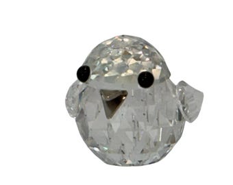 May include: A clear, faceted crystal bird ornament with black eyes and a silver beak. The bird has small wings and a rounded body. The crystal reflects light, creating a sparkling effect. The ornament is a decorative item.
