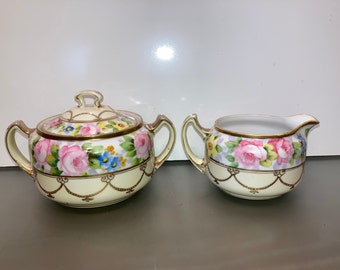 May include: A vintage porcelain sugar and creamer set with floral designs. The set features pink roses, blue and yellow flowers, and gold accents. The creamer has a handle and a spout, while the sugar bowl has a lid.