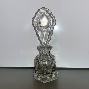 Puede incluir: Un frasco de perfume de vidrio transparente con un tapón decorativo y adornado. El frasco tiene un diseño texturizado y facetado y una base redonda.