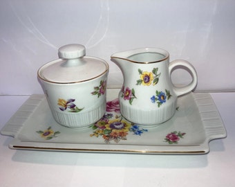 May include: A white porcelain sugar bowl and creamer set with a floral design. The set is on a matching tray with a gold trim.
