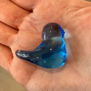 May include: A small, clear blue glass bird figurine. The bird is sitting with its head up and beak slightly open. The glass is a deep, vibrant blue color, with a smooth, glossy finish. The figurine is approximately 3 inches long.