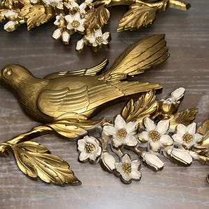 Pode incluir: Escultura de p&aacute;ssaro em metal dourado pousada em um galho com flores e folhas brancas e douradas. A pe&ccedil;a de arte decorativa de parede tem um design detalhado e &eacute; adequada para decora&ccedil;&atilde;o de casa.