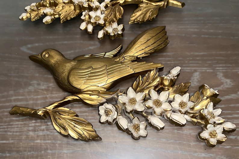 Pode incluir: Escultura de p&aacute;ssaro em metal dourado pousada em um galho com flores e folhas brancas e douradas. A pe&ccedil;a de arte decorativa de parede tem um design detalhado e &eacute; adequada para decora&ccedil;&atilde;o de casa.