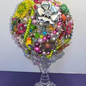 May include: A colorful, round bouquet of faux flowers made from vintage jewelry and beads. The bouquet is displayed on a clear glass pedestal.
