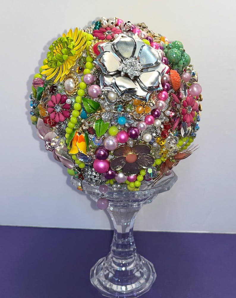 May include: A colorful, round bouquet of faux flowers made from vintage jewelry and beads. The bouquet is displayed on a clear glass pedestal.