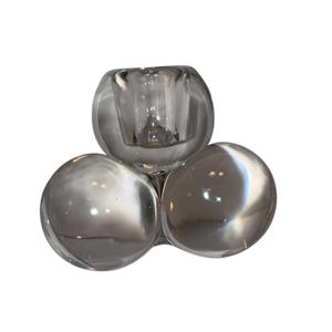 May include: A clear glass candle holder with a unique design. The holder features three rounded, connected sections, with a central cup for a candle. The glass has a smooth, reflective surface, creating a modern aesthetic.