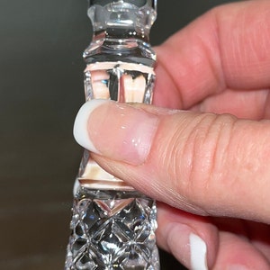 May include: A clear, faceted crystal bottle stopper with a geometric design. The stopper has a hexagonal top and a diamond-patterned body. The object is held by a hand with a white-tipped manicure.