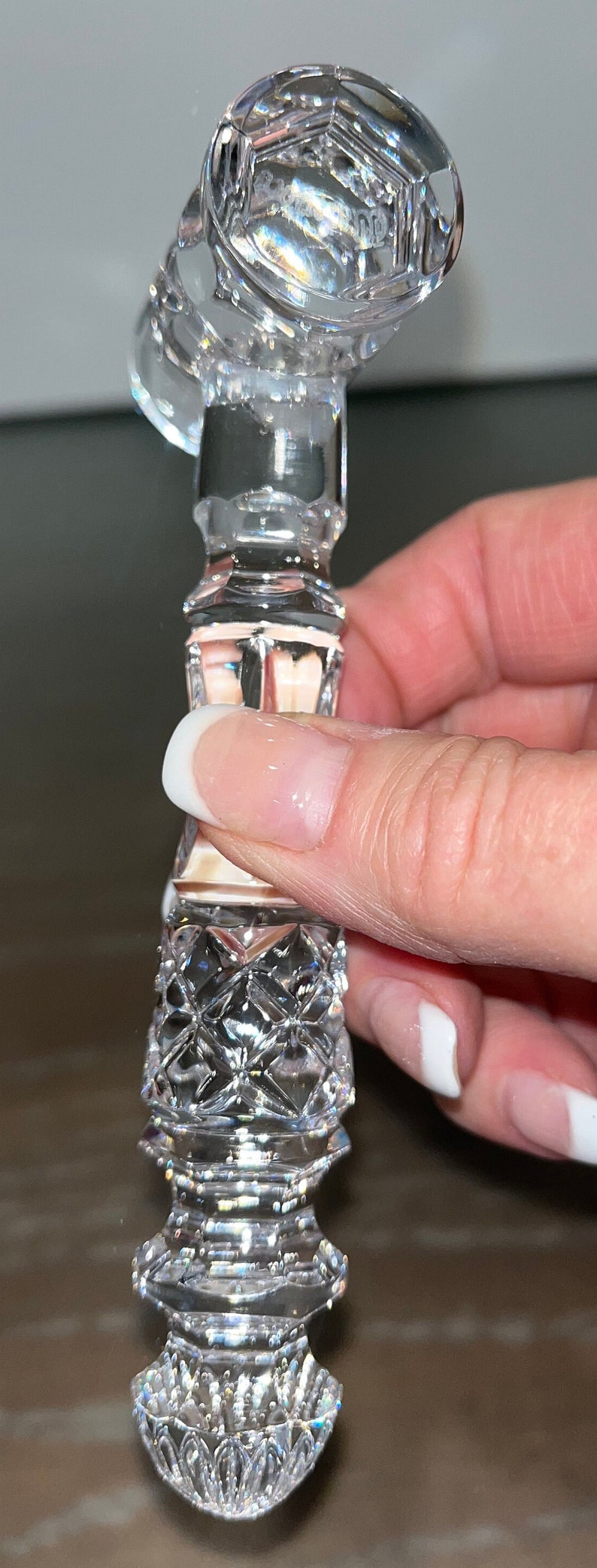 May include: A clear, faceted crystal bottle stopper with a geometric design. The stopper has a hexagonal top and a diamond-patterned body. The object is held by a hand with a white-tipped manicure.