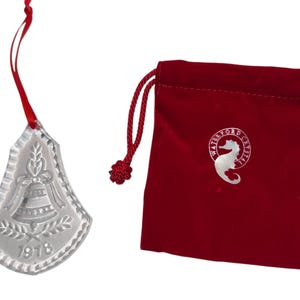 Op de afbeelding: Een helder kristallen belvormig ornament met een rood lint, met een reliëfklok, bladeren en het jaartal 1978. Een rode fluwelen zakje met een zeepaardlogo staat naast het ornament.