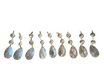 May include: Eight clear crystal chandelier pendants with gold-colored connectors. Each pendant features a teardrop-shaped crystal hanging below three smaller, round crystals. The crystals reflect light.