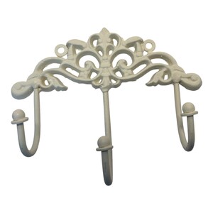 May include: A cream-colored wall-mounted hook rack featuring three individual hooks with rounded tips. The top section displays an ornate, symmetrical scrollwork design with two circular mounting holes integrated into the metal frame structure.
