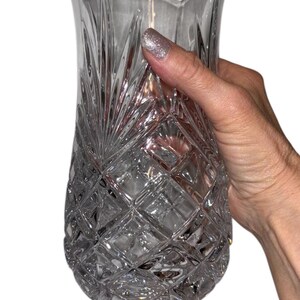May include: Clear crystal vase with a diamond-cut pattern and a scalloped rim. The vase is held by a hand with glittery nail polish. The vase has a wide base and a flared top, suitable for floral arrangements.