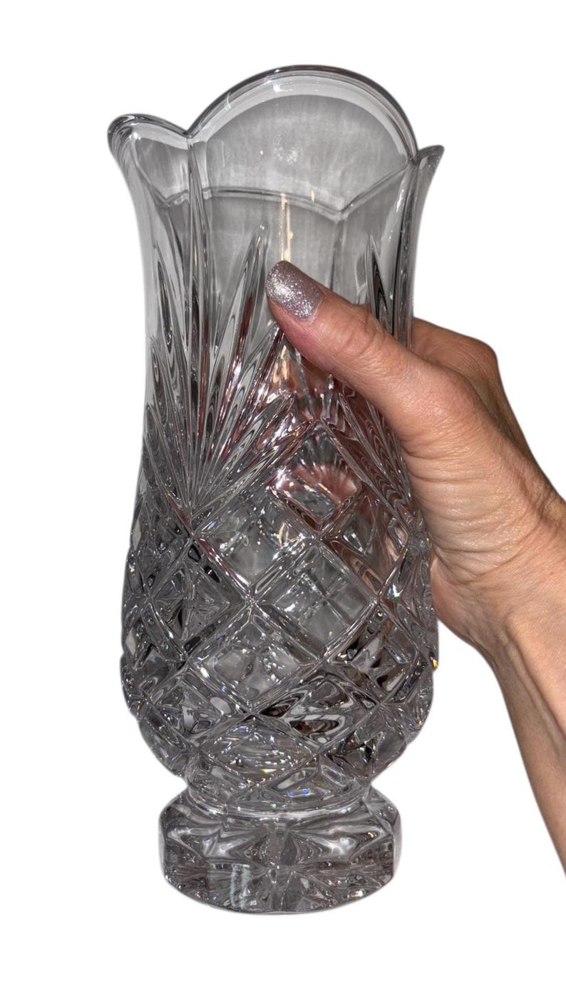 May include: Clear crystal vase with a diamond-cut pattern and a scalloped rim. The vase is held by a hand with glittery nail polish. The vase has a wide base and a flared top, suitable for floral arrangements.