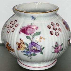 May include: A white porcelain vase with a round, ribbed body and a floral design. The vase has a gold rim and a red and white base. There are small holes around the body of the vase.