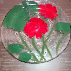 Puede incluir: Un plato de vidrio transparente con un diseño floral. El plato presenta dos flores rojas con hojas verdes.