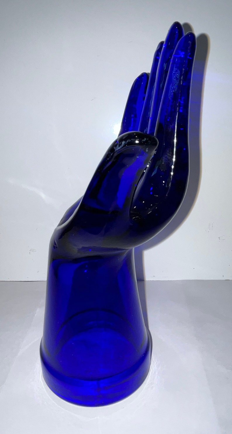 Peut inclure: Une sculpture de main en verre bleu cobalt. La main est ouverte et les doigts sont tendus. La sculpture est faite de verre &eacute;pais et a une finition lisse et polie.