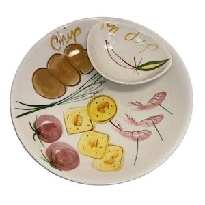May include: A white ceramic chip and dip serving platter with hand-painted illustrations of food items. The platter features the words "chip" and "in dip" in gold lettering. The illustrations include chips, cheese, shrimp, and tomatoes.