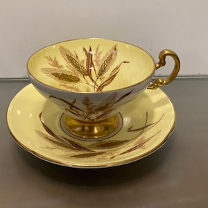 Puede incluir: Un juego de taza y platillo de té amarillo con borde dorado y diseño de trigo marrón.