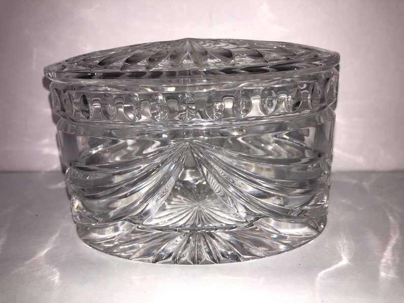 May include: A clear glass oval-shaped trinket box with a lid. The box has a cut glass design with a starburst pattern on the bottom and a decorative band around the top.