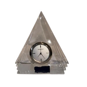 May include: A clear, pyramid-shaped clock with a silver-colored clock face. The clock face has black hands and the word "MARQUIS" printed on it. The clock is made of a clear, possibly glass, material.