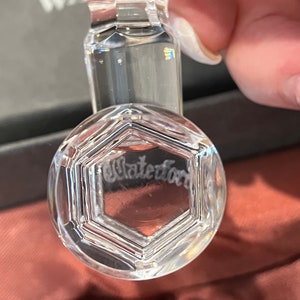 May include: A clear crystal bottle stopper with a hexagonal design and the etched word "Waterford". The stopper has a cylindrical stem and a rounded top, reflecting light. The background is a blurred, neutral color.