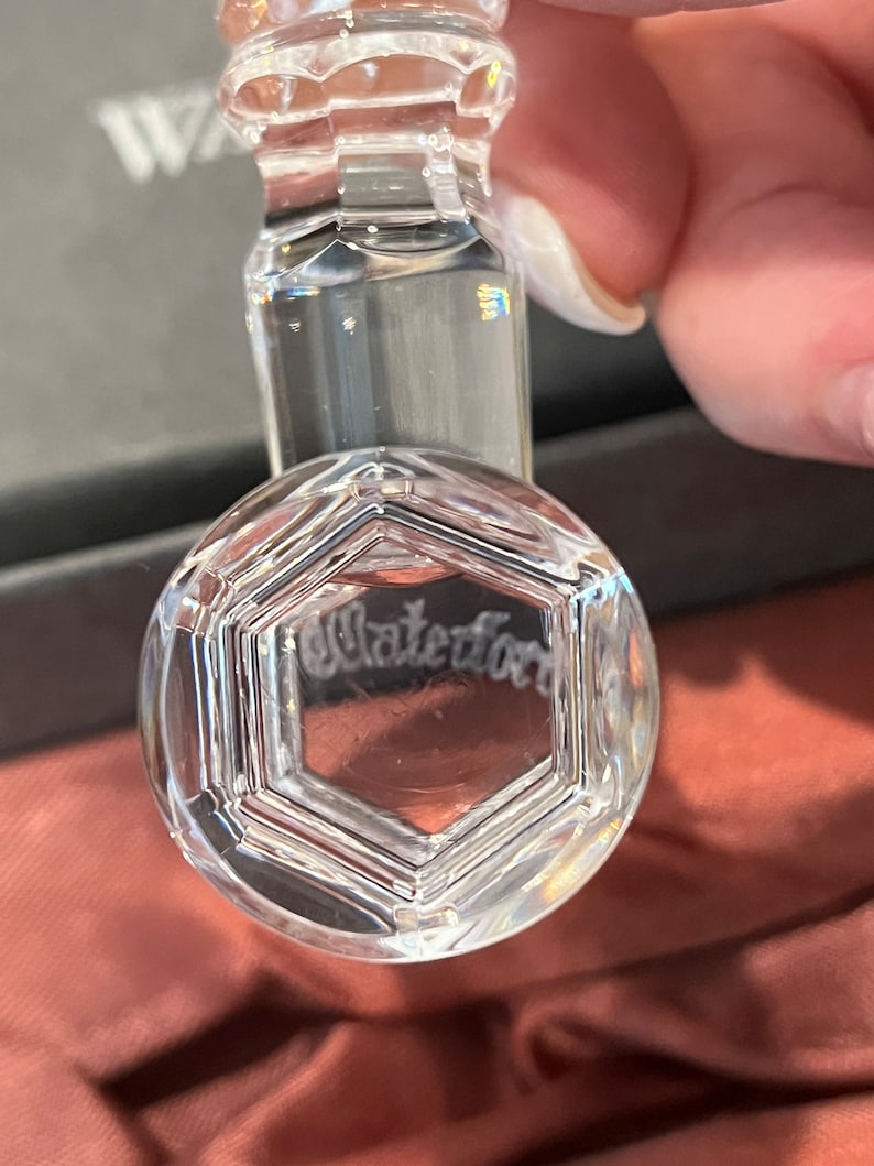May include: A clear crystal bottle stopper with a hexagonal design and the etched word "Waterford". The stopper has a cylindrical stem and a rounded top, reflecting light. The background is a blurred, neutral color.