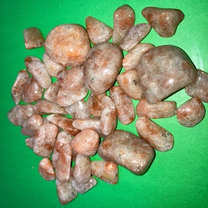 May include: A collection of tumbled sunstone crystals in various sizes and shapes. The stones are a light orange color with a shimmery, iridescent finish.